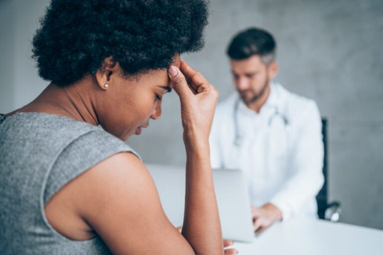 African-american-woman-sitting-opposite-the-doctor-in-his-office-and-holding-her-head-with-hand.