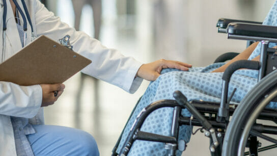 Individual_with_disability_sits_in_wheelchair_as_doctor_approaches_with_hand_on_their_knee_in_empathetic_manner.