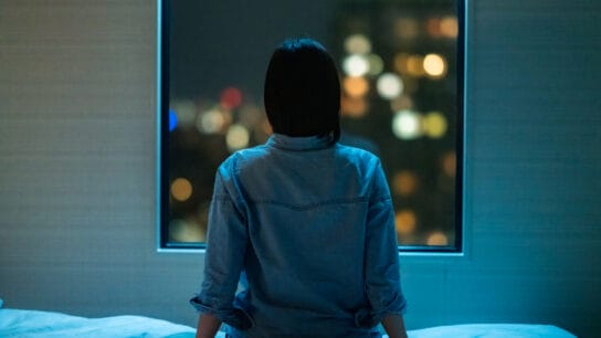 A-rear-view-of-a- woman-sitting-alone -on-a-bed-in-room- and-looking-through-the -window-at-night.