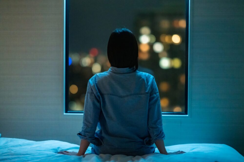 A-rear-view-of-a- woman-sitting-alone -on-a-bed-in-room- and-looking-through-the -window-at-night.
