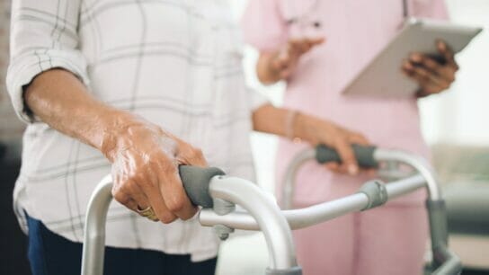 Cropped_image_of_an_ unrecognizable_older_ woman_using_a_walker_ and_walking_with_the_ assistance_of_a_ physiotherapist.