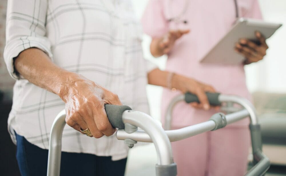 Cropped_image_of_an_ unrecognizable_older_ woman_using_a_walker_ and_walking_with_the_ assistance_of_a_ physiotherapist.